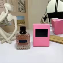 Small bottle of perfume next to a pink box on a surface with a mirror and sculpture in the background.